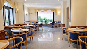 A hotel dining area with round tables and blue chairs, large windows with curtains, and a view of greenery outside. - Phi Hotel Ambra (Pescara)