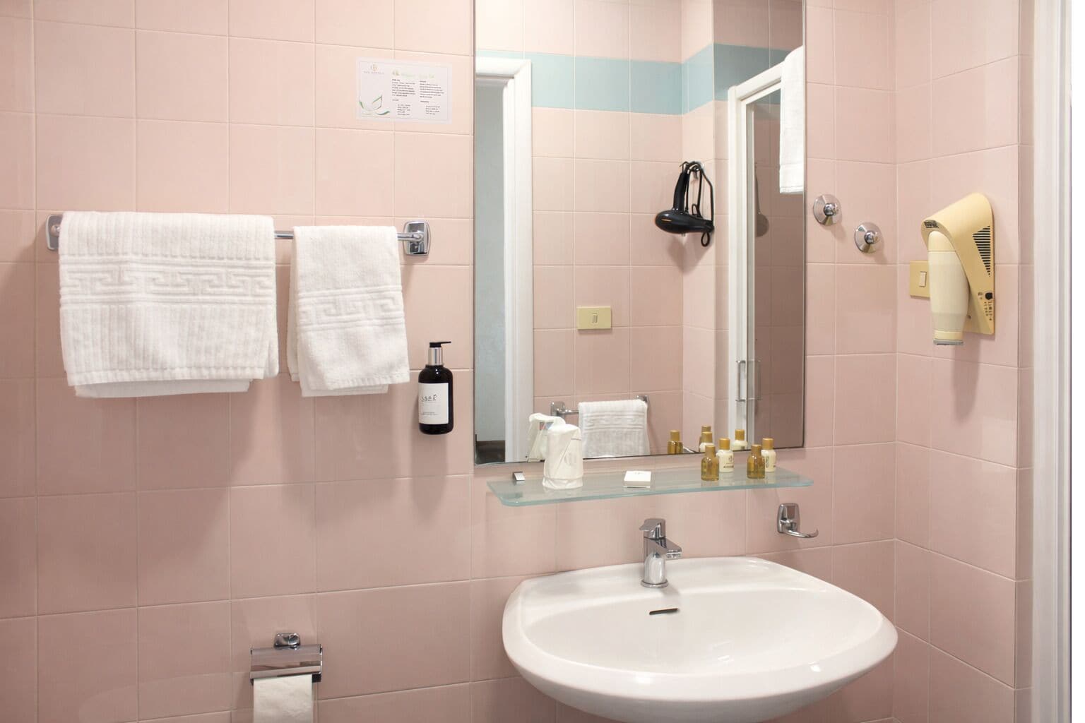 A bathroom with pink tiles, a white sink, two white towels, a mirror, a hairdryer, and a bottle of lotion.
