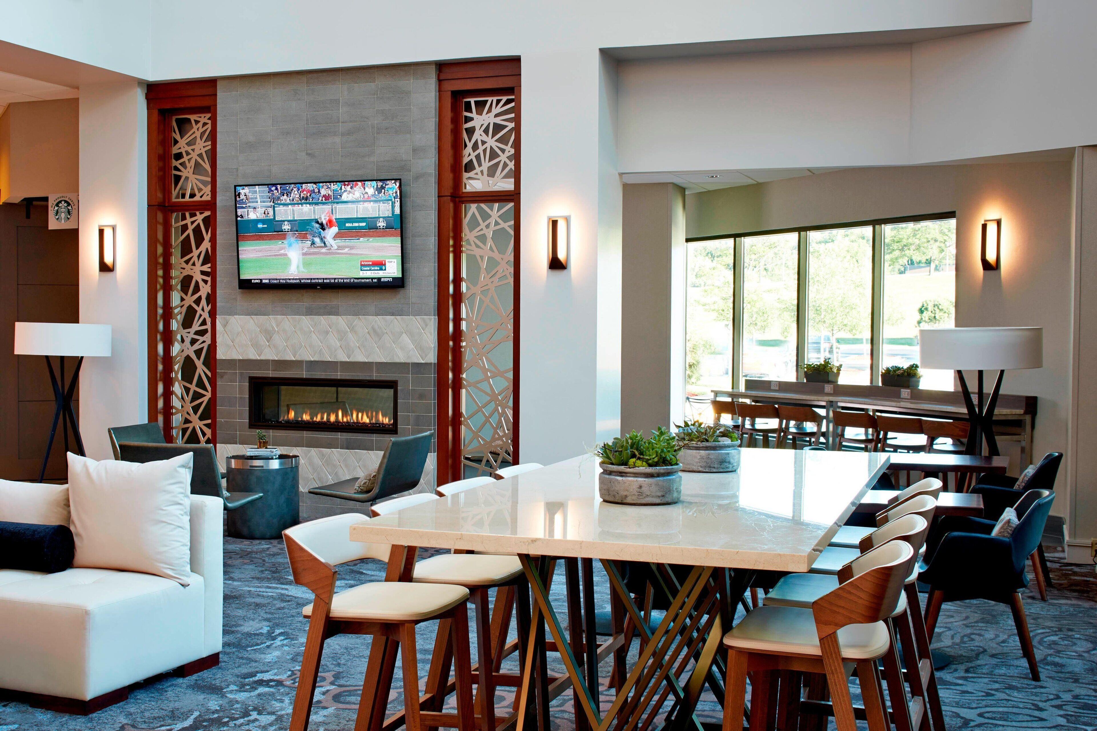 A modern hotel lobby with a fireplace, a large TV screen, comfortable seating, and a dining area.