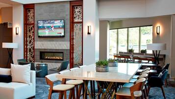 A modern hotel lobby with a fireplace, a large TV screen, comfortable seating, and a dining area.