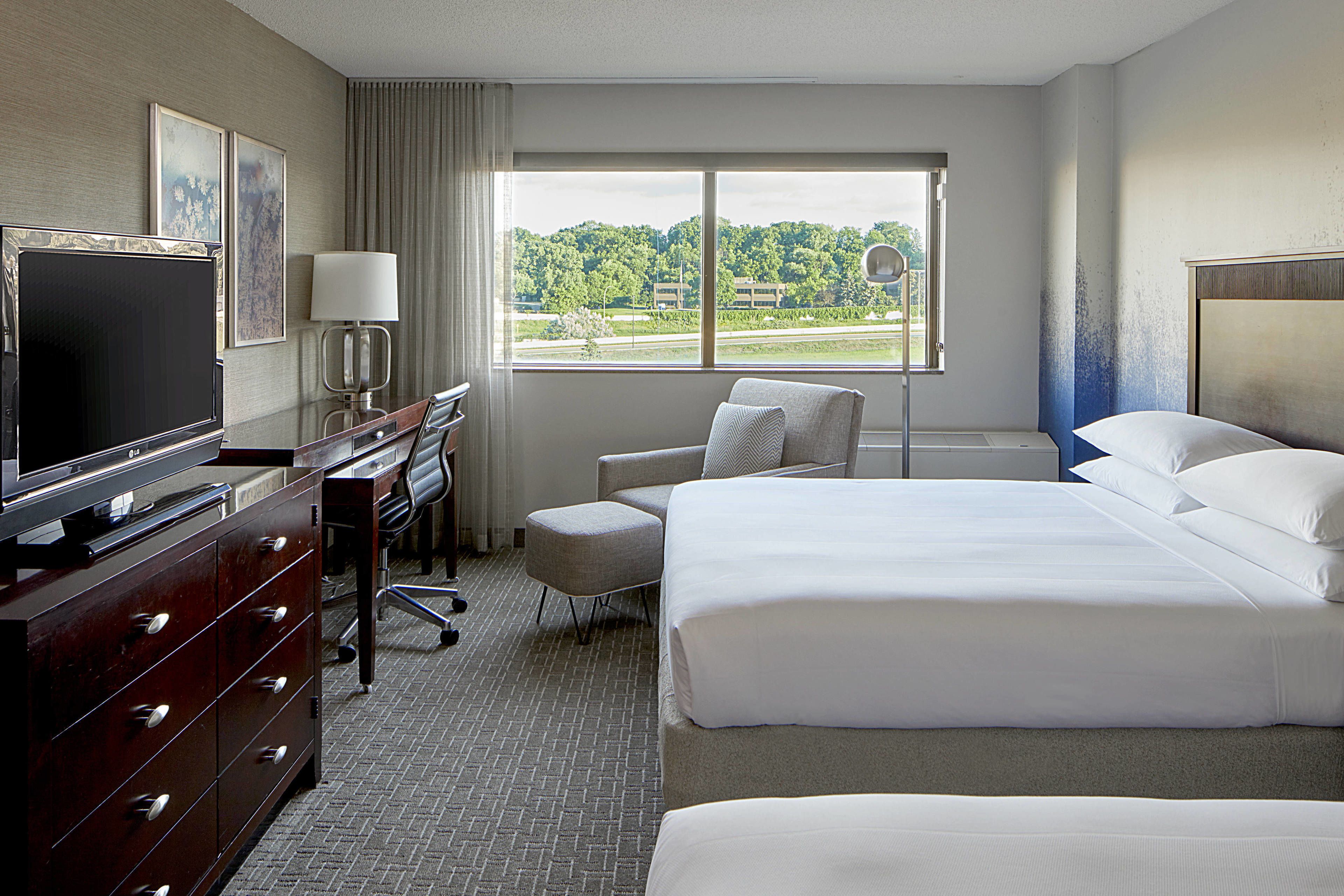 A hotel room with a large bed, a desk with a TV, a chair, and a window with a view of greenery.
