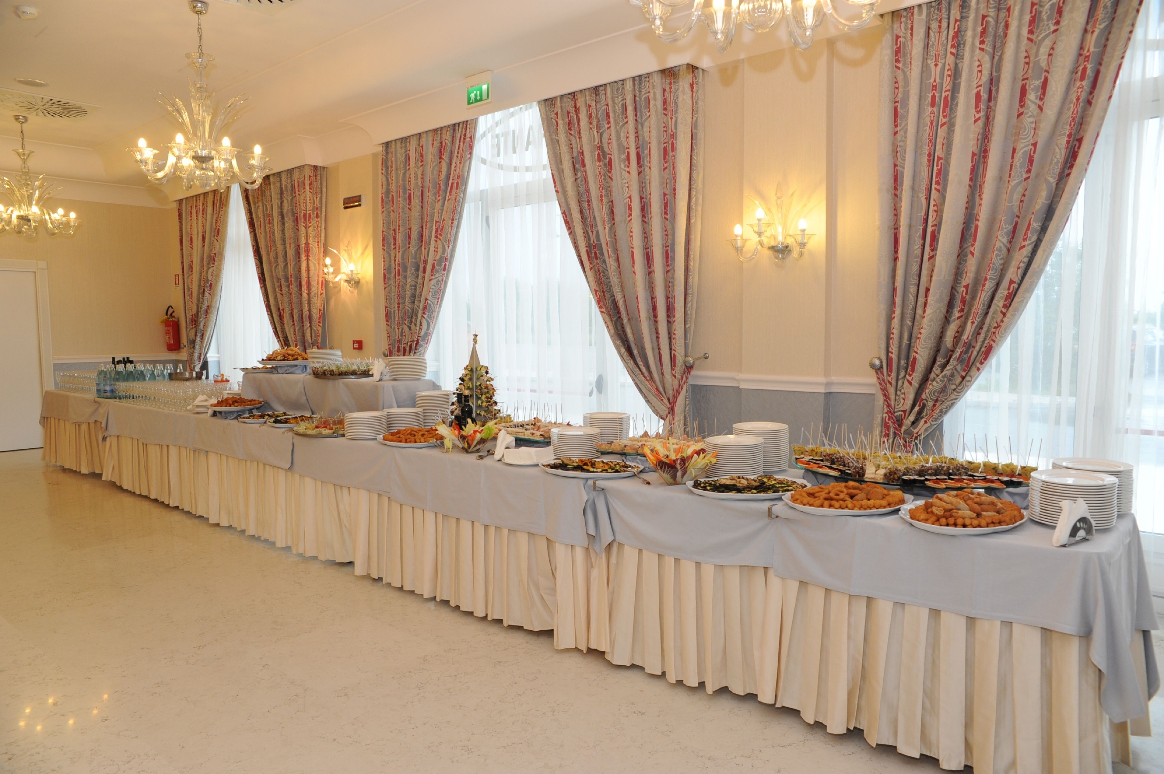Ein reich gedeckter Buffet mit verschiedenen Speisen und eine kleine Weihnachtstanne, platziert in einem Raum mit Kronleuchtern und gemusterten Vorhängen.