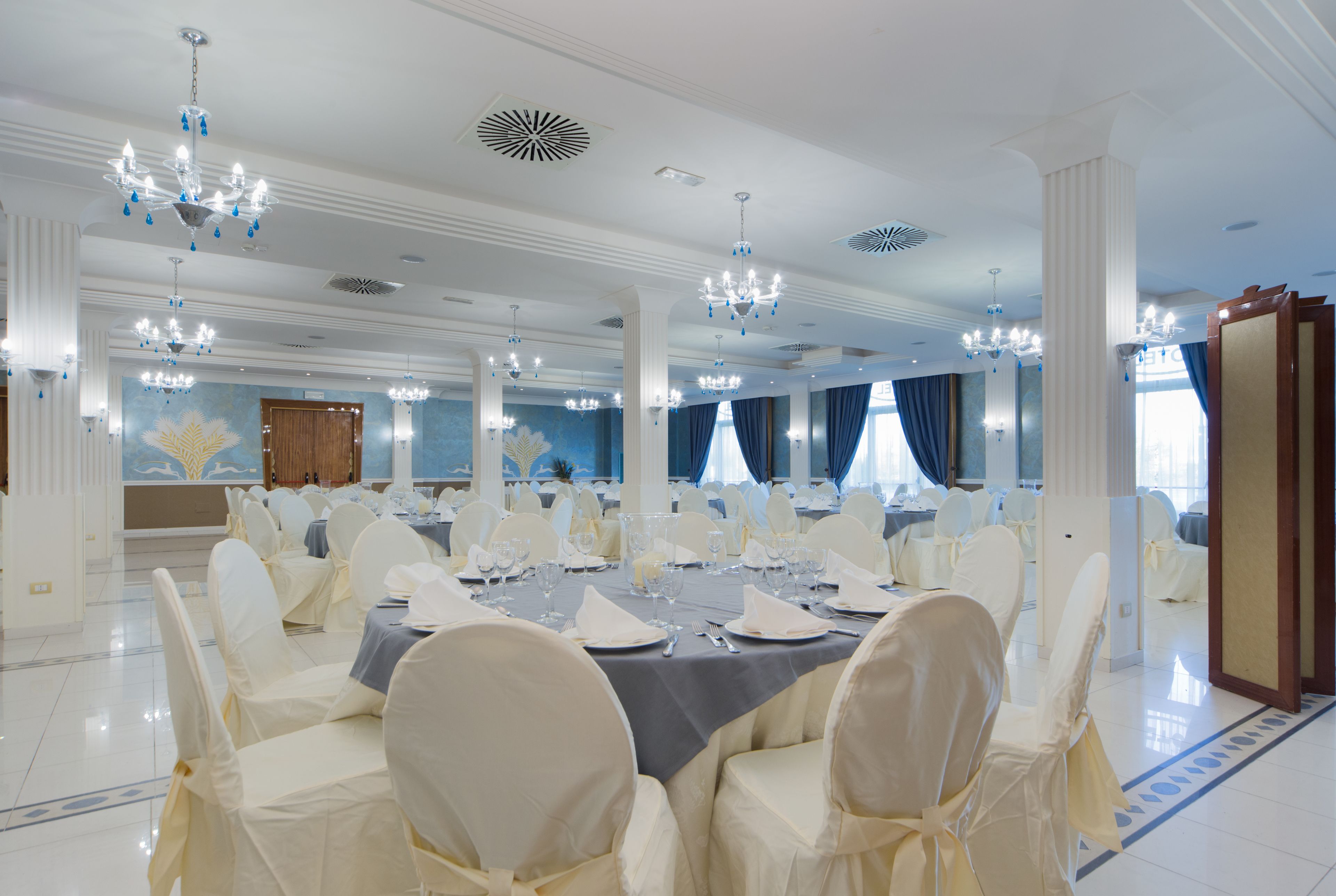A banquet hall with round tables, white chairs, chandeliers, and blue curtains.