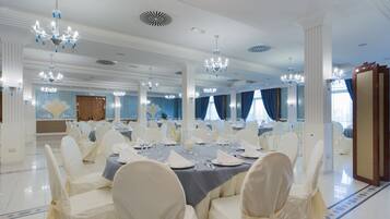 A banquet hall with round tables, white chairs, chandeliers, and blue curtains.