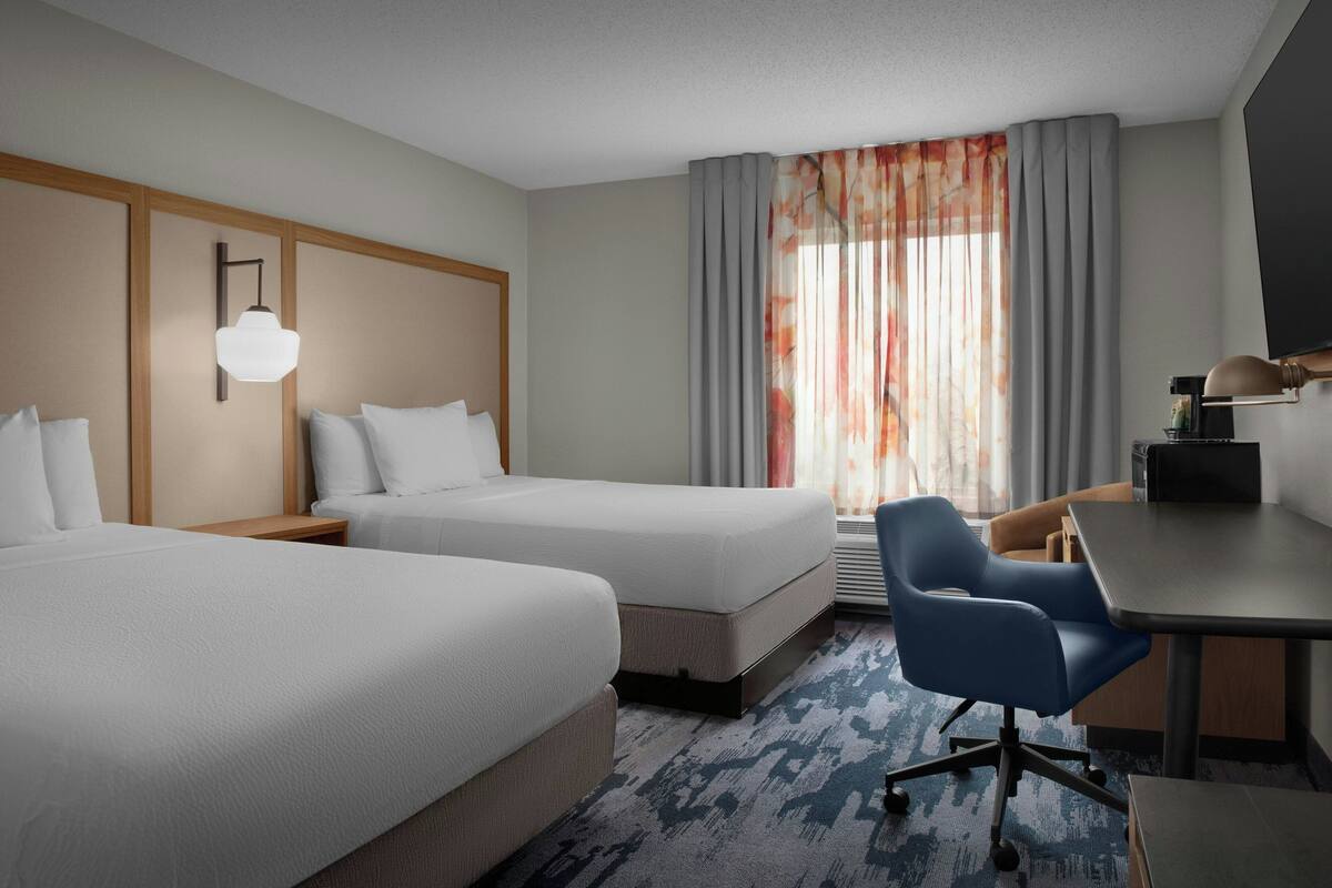 A hotel room with two beds, a desk, a chair, and a large window with curtains.