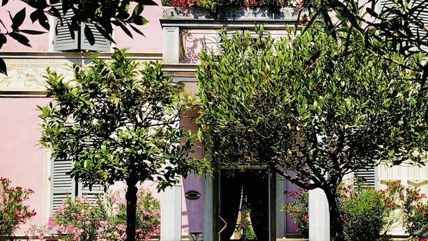 Un chemin mène à un bâtiment à la façade rose, aux volets blancs et à une entrée centrale surmontée d’un arc décoratif.