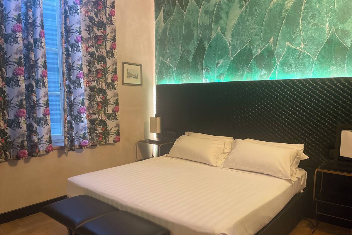 Habitación de hotel con cama, banca, mesita de noche con lámpara y pared con diseño de hojas.