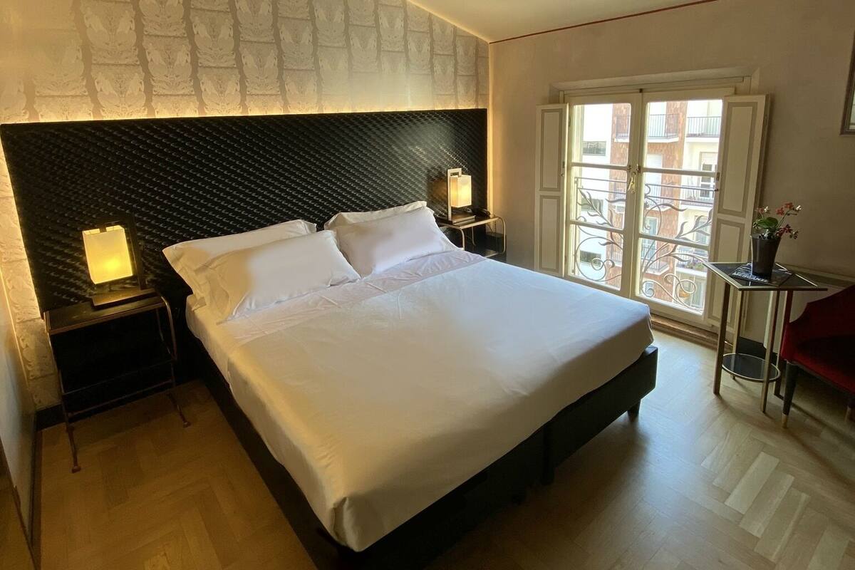 Habitación de hotel con una cama grande, una mesita de noche con lámpara, una pequeña mesa con una planta y vista al balcón exterior a través de una ventana.