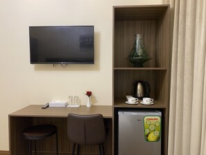 Quarto de hotel com escrivaninha de madeira, televisão, frigorífico, máquina de café e um vaso com uma flor.
