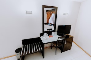 Quarto pequeno e arrumado, com escrivaninha, cadeira e televisão. Um espelho na parede reflete uma janela com cortinas.