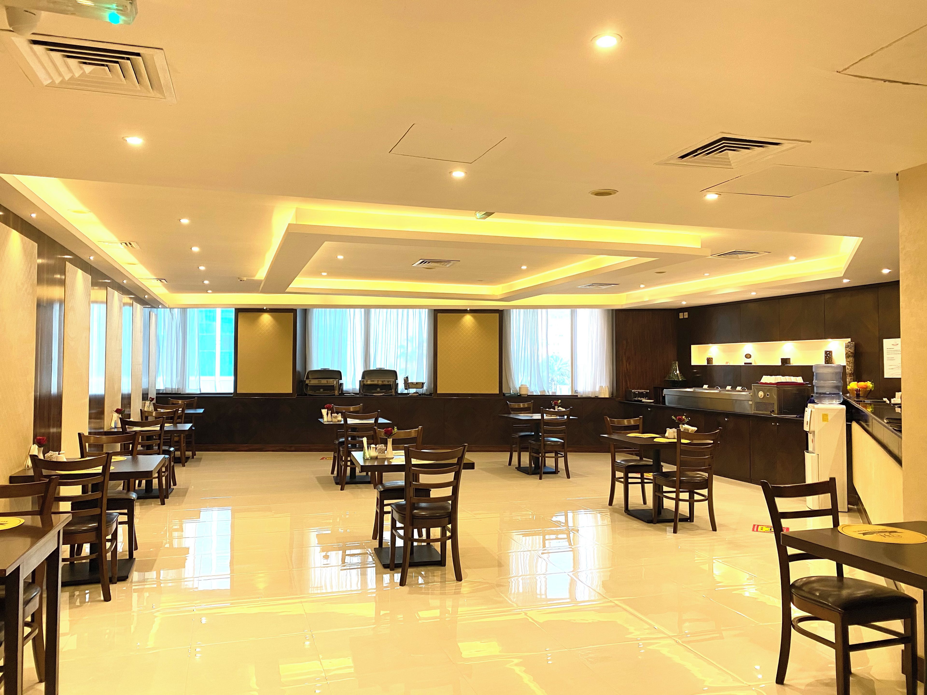 Une salle à manger d’hôtel avec de nombreuses tables et chaises, un bar et de grandes fenêtres.