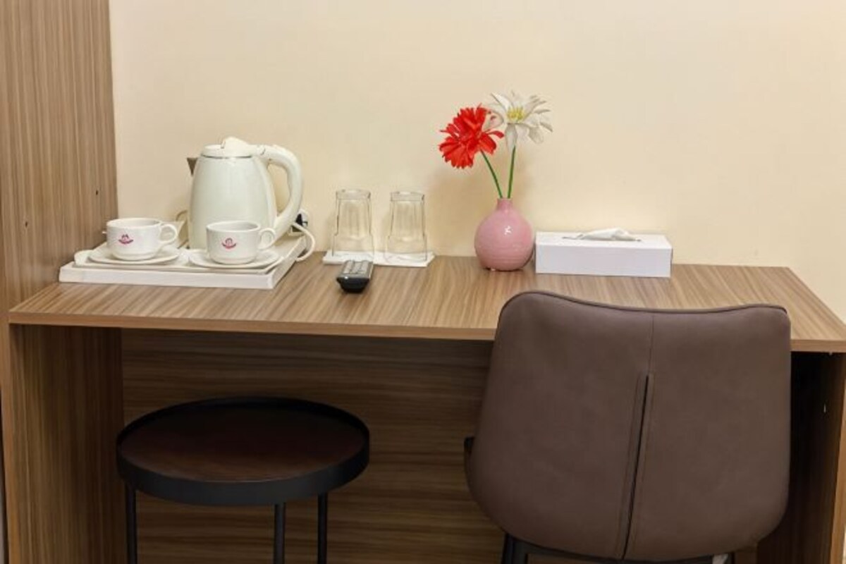 Een houten bureau met een witte theepot, kopjes, glazen en een vaas met bloemen. Er staat een bruine stoel en een zwarte kruk.