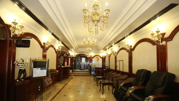 Um saguão de hotel com piso de mármore, balcão de recepção de madeira, uma estátua, cadeiras de massagem e um lustre.