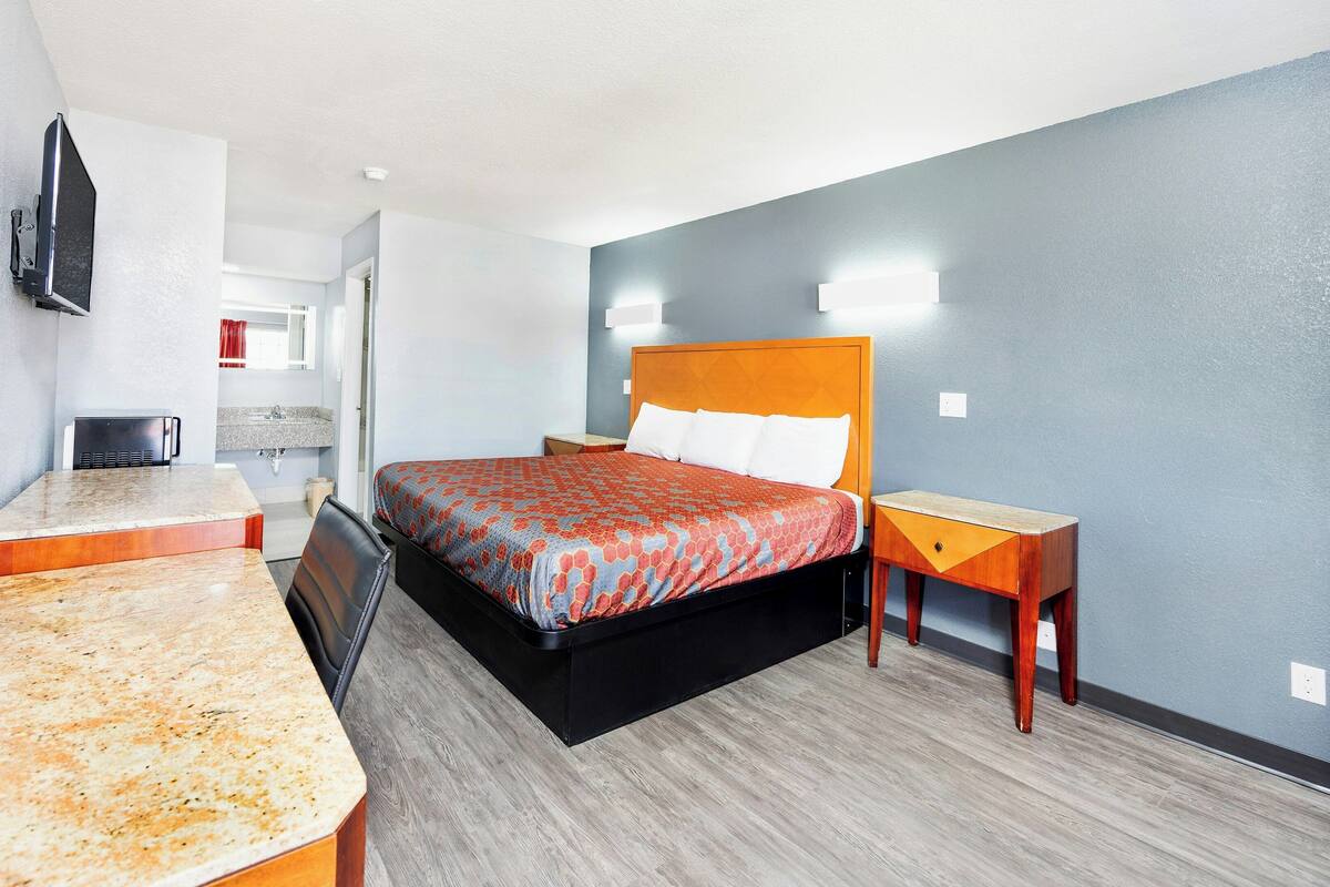 A hotel room with a bed, a desk, a chair, a telephone, a television, and a bathroom visible through an open door.