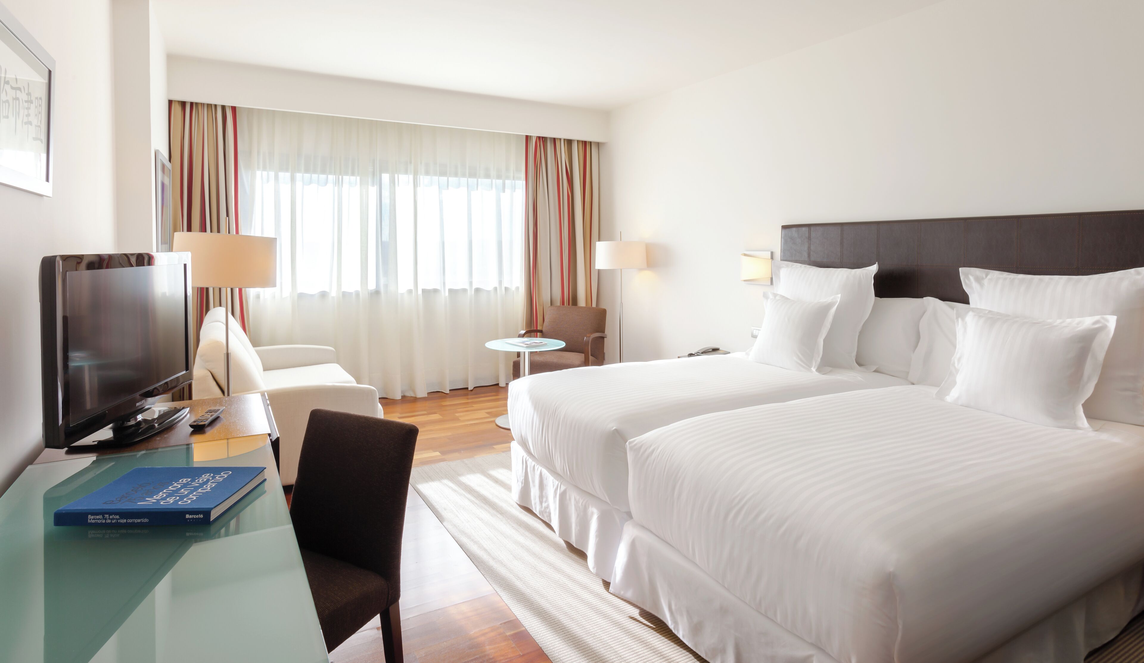 Hotellihuone, jossa on kaksi sänkyä, televisio, työpöytä lampun kanssa, tuoli ja pieni pöytä.