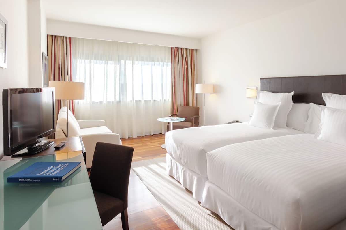 Hotellihuone, jossa on kaksi sänkyä, televisio, työpöytä lampun kanssa, tuoli ja pieni pöytä.