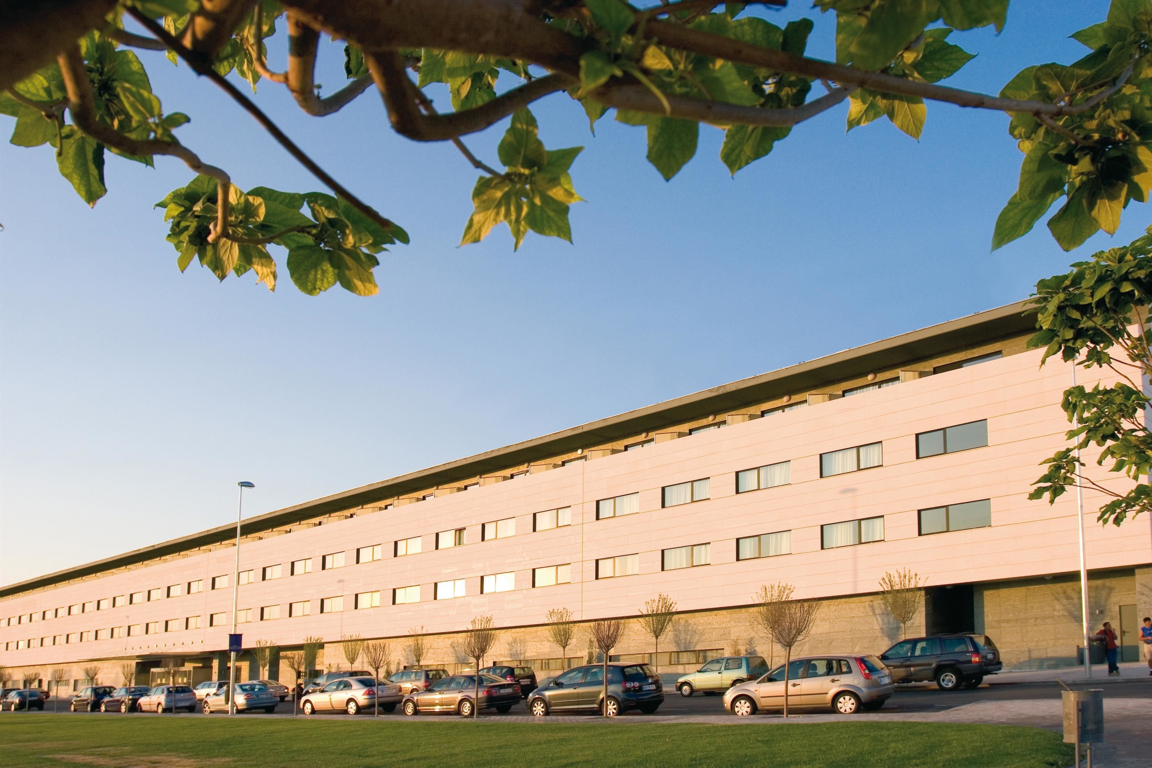 A modern building with a flat roof and multiple windows. There are several cars parked in front of the building.