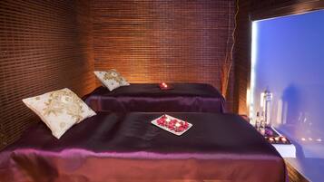 Ein Zimmer mit zwei Betten, jeweils mit lila Bettwäsche und einem Zierkissen. Auf einem Bett steht ein Tablett mit roten Rosenblättern und einer Kerze. Das Zimmer hat eine Backsteinwand und ein großes Fenster mit Blick auf die Stadt.
