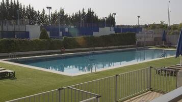 Una piscina rectangular con un borde pavimentado, una valla metálica y una zona verde con algunas sillas de descanso.