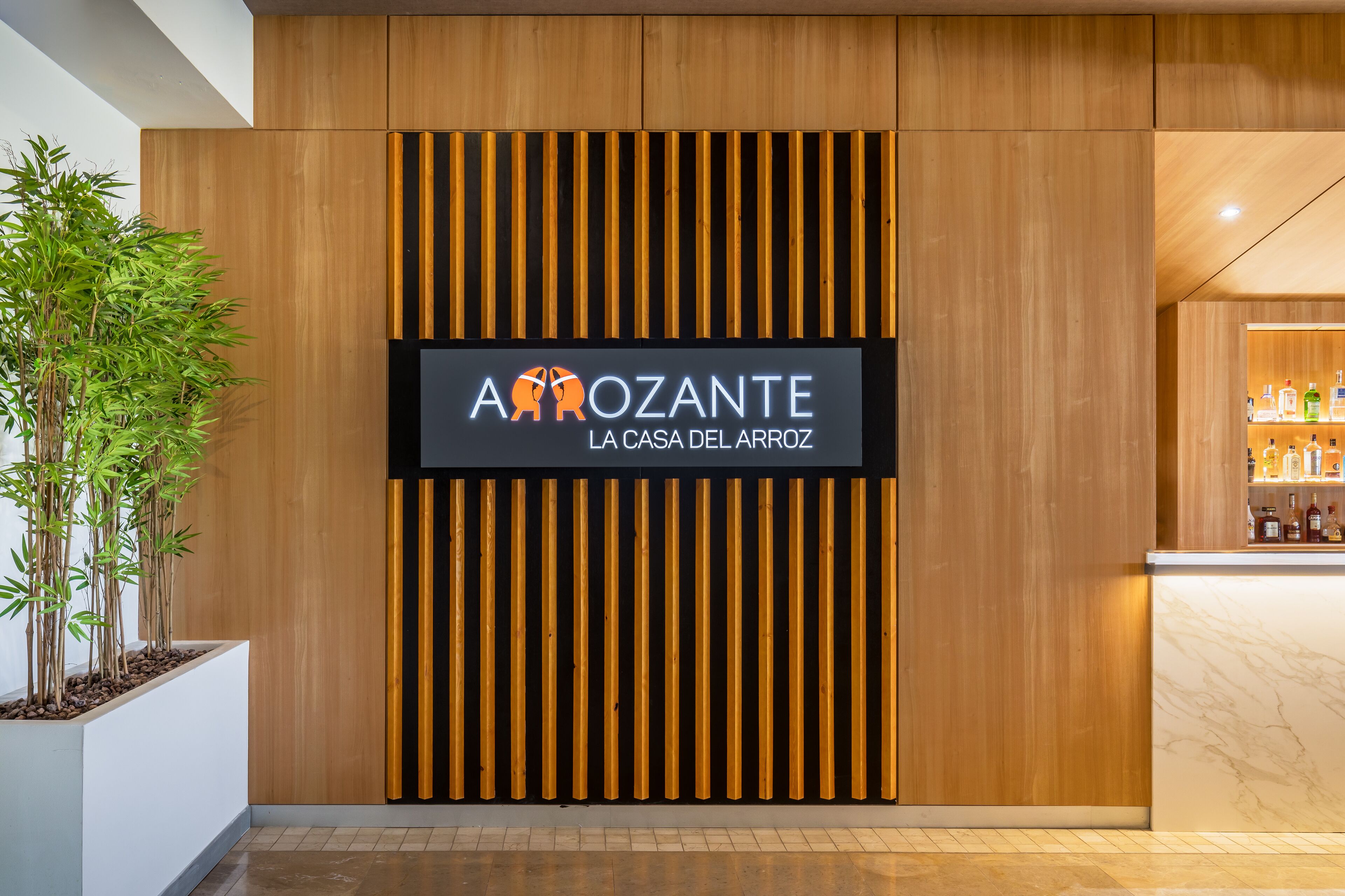 Ein modernes Interieur mit einer Wandpaneele aus Holz, einem Schild mit der Aufschrift 'AroZANTE LA CASA DEL ARROZ' und einer Bar mit Flaschen.