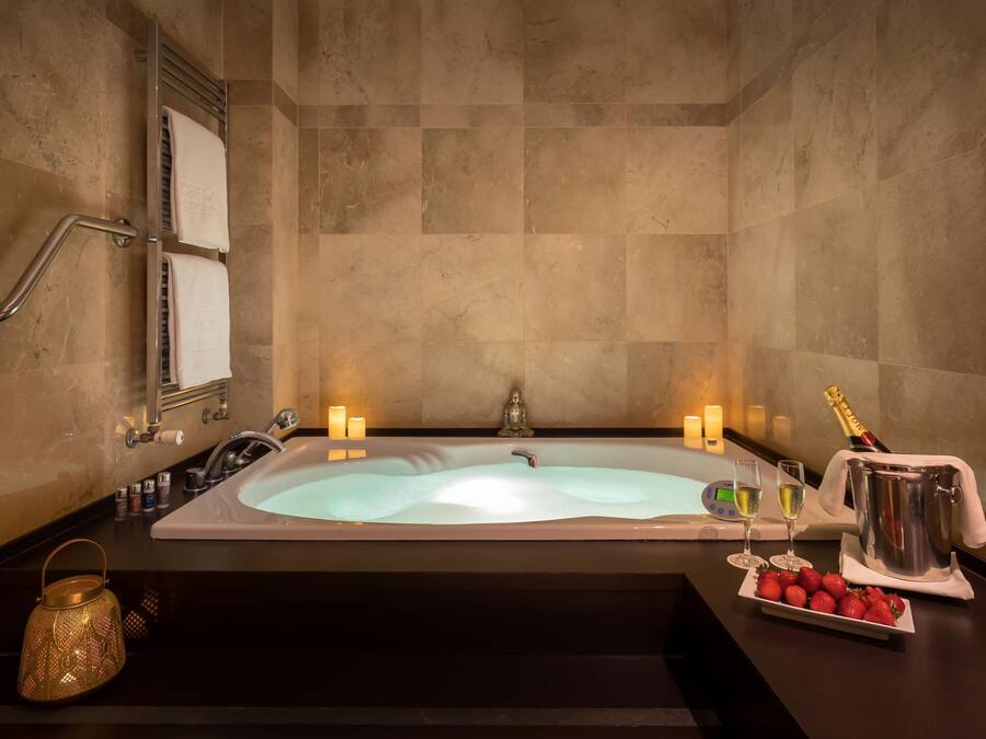 Une salle de bain luxueuse avec une grande baignoire, une barre d’appui, un porte-serviettes, des bougies, une bouteille de champagne et un plateau de fraises.