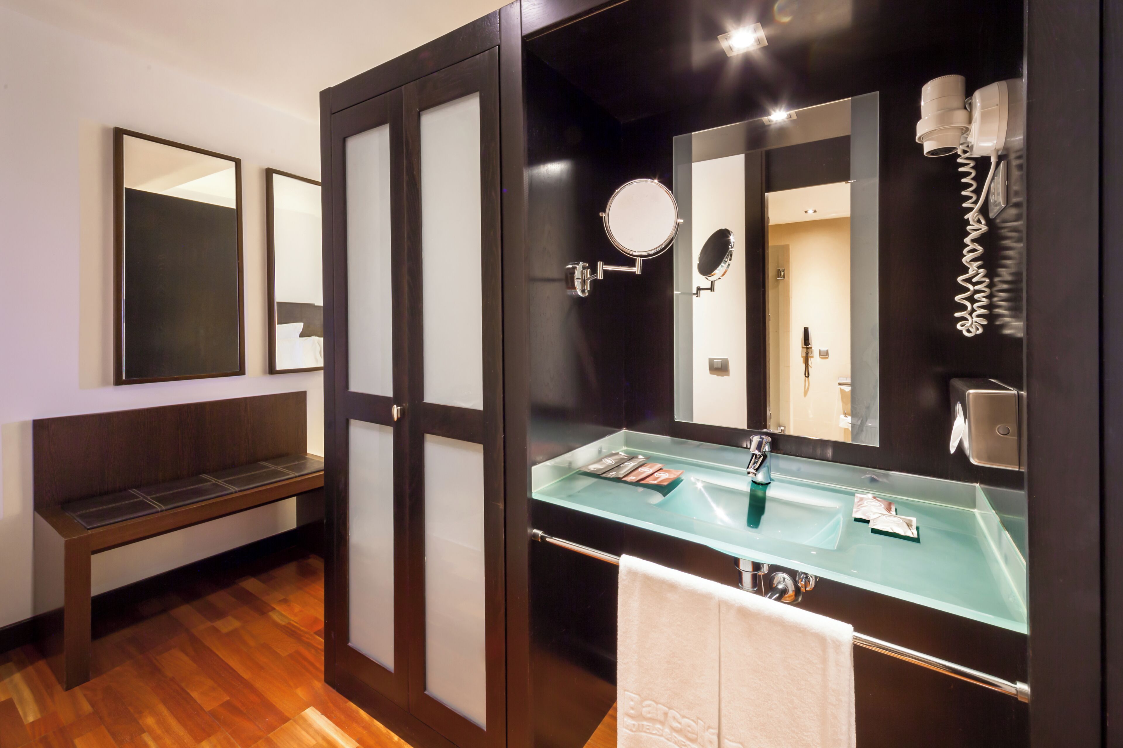 A bathroom with a glass sink, a towel rack, a bench, and a mirror with a round mirror and a hairdryer.