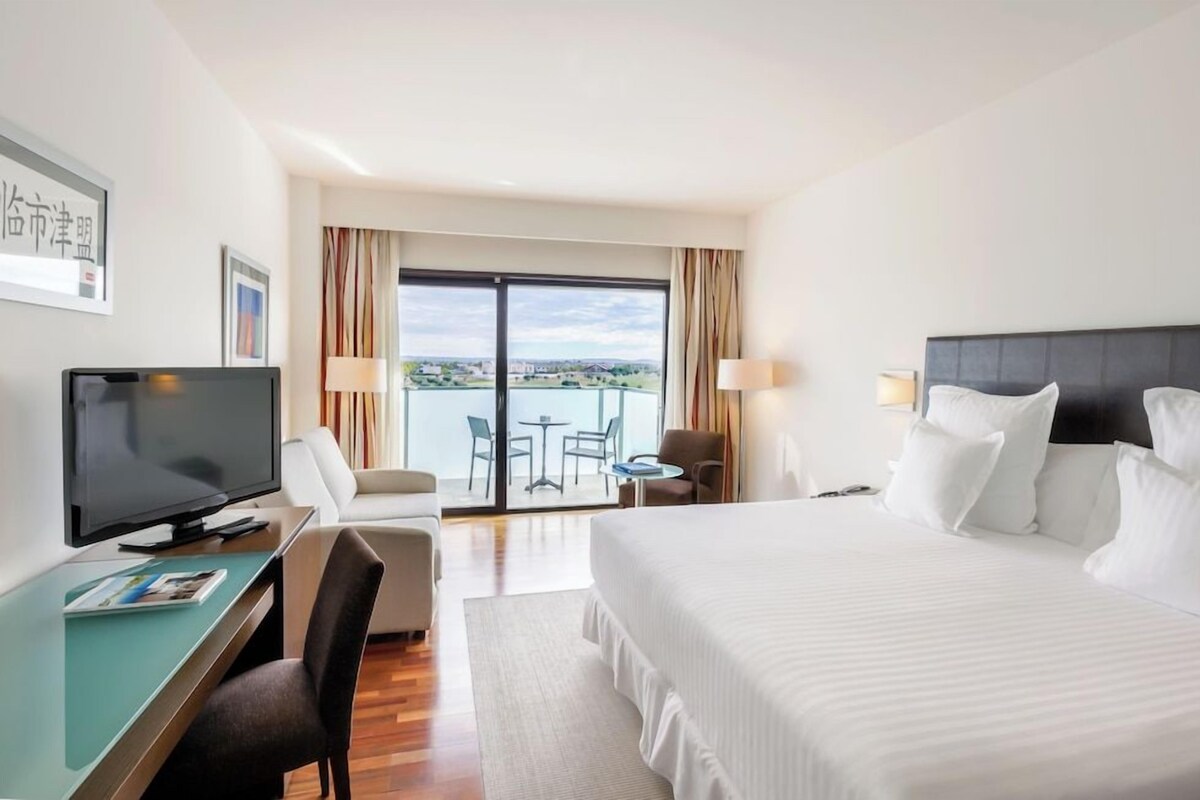 Une chambre d’hôtel comprenant un lit, un bureau avec une télévision, une chaise, une petite table et une vue sur l’extérieur grâce à une grande fenêtre.