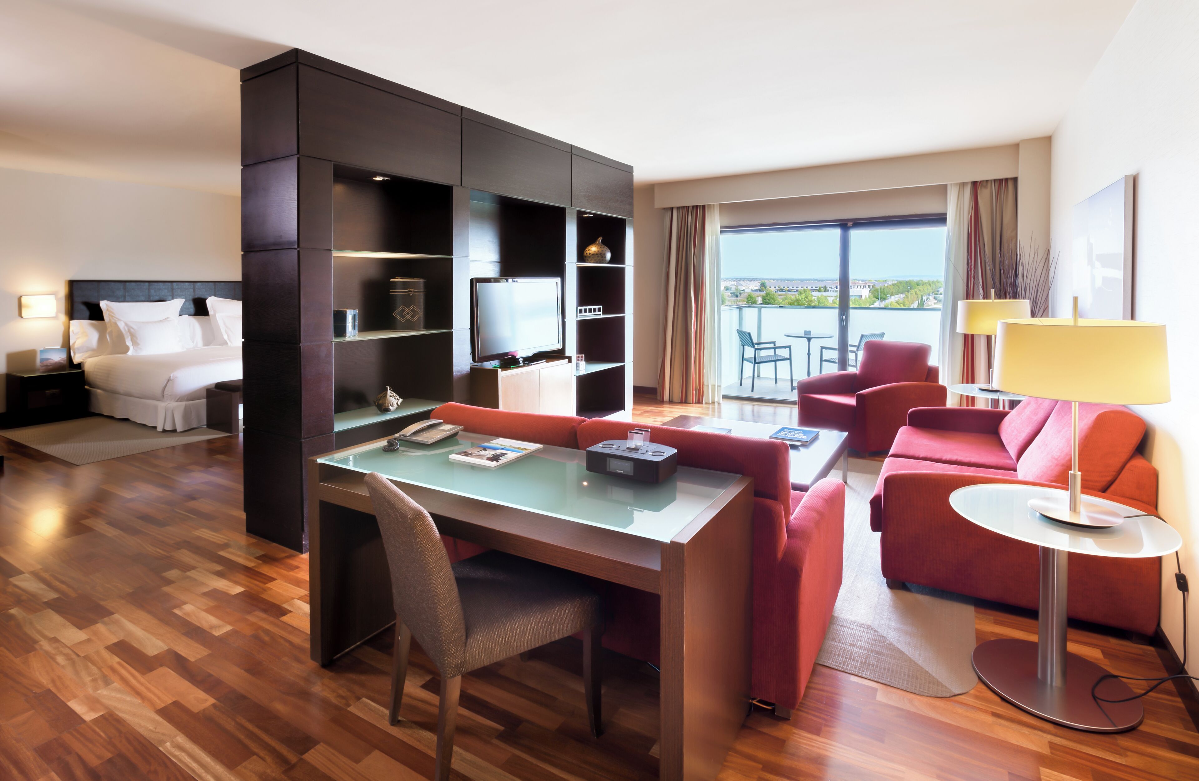 A modern hotel room with a dining area, a sofa, a red armchair, a small table with a lamp, and a large window with a view.