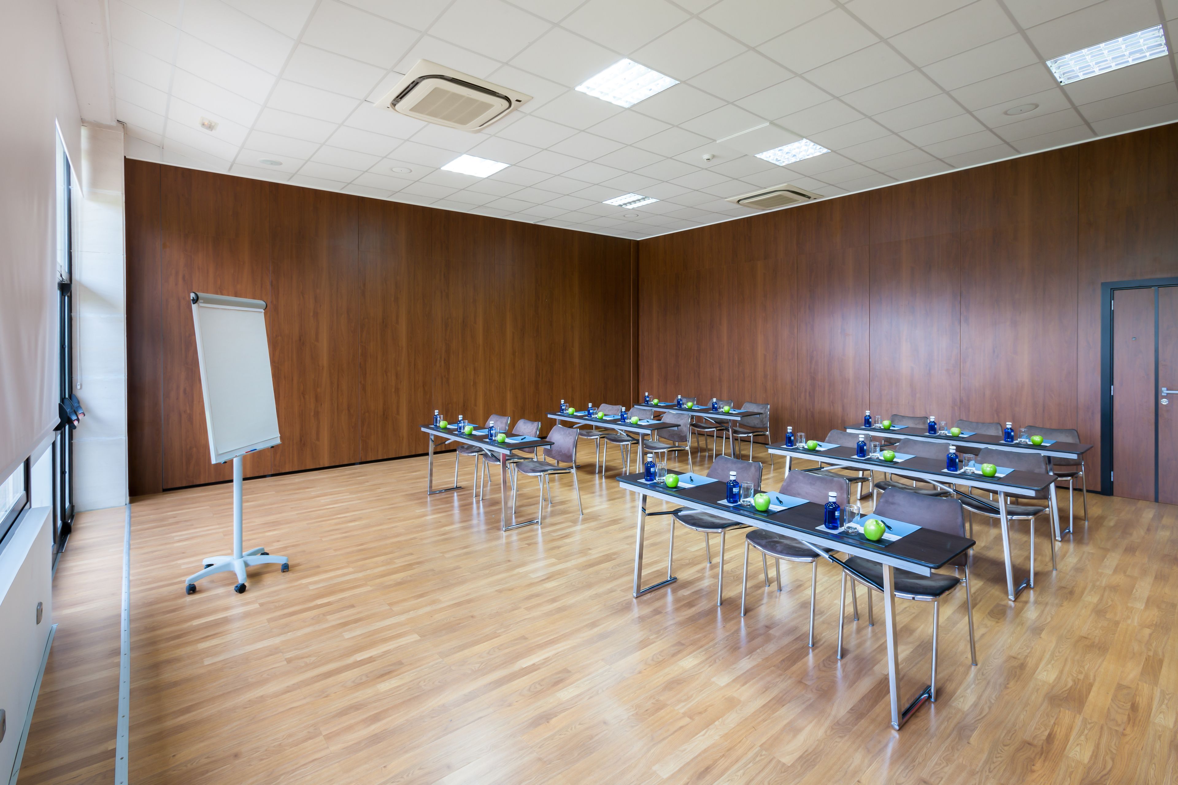 Un salón de conferencias con paredes de madera, una pizarra blanca y filas de mesas con sillas y botellas de agua.