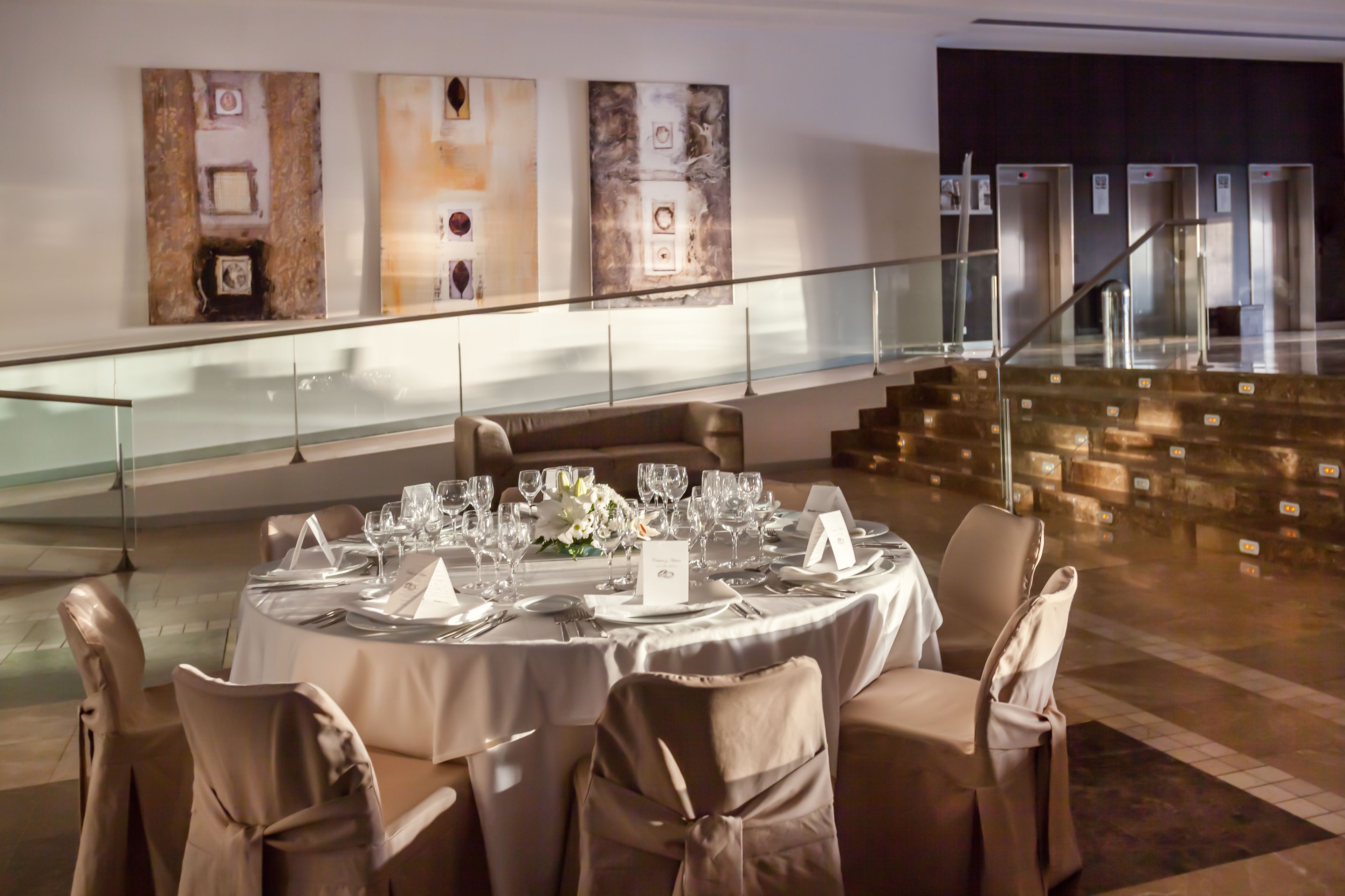 Une salle à manger avec des tables rondes dressées pour un repas, des chaises ornées de nœuds papillon et un escalier en arrière-plan.