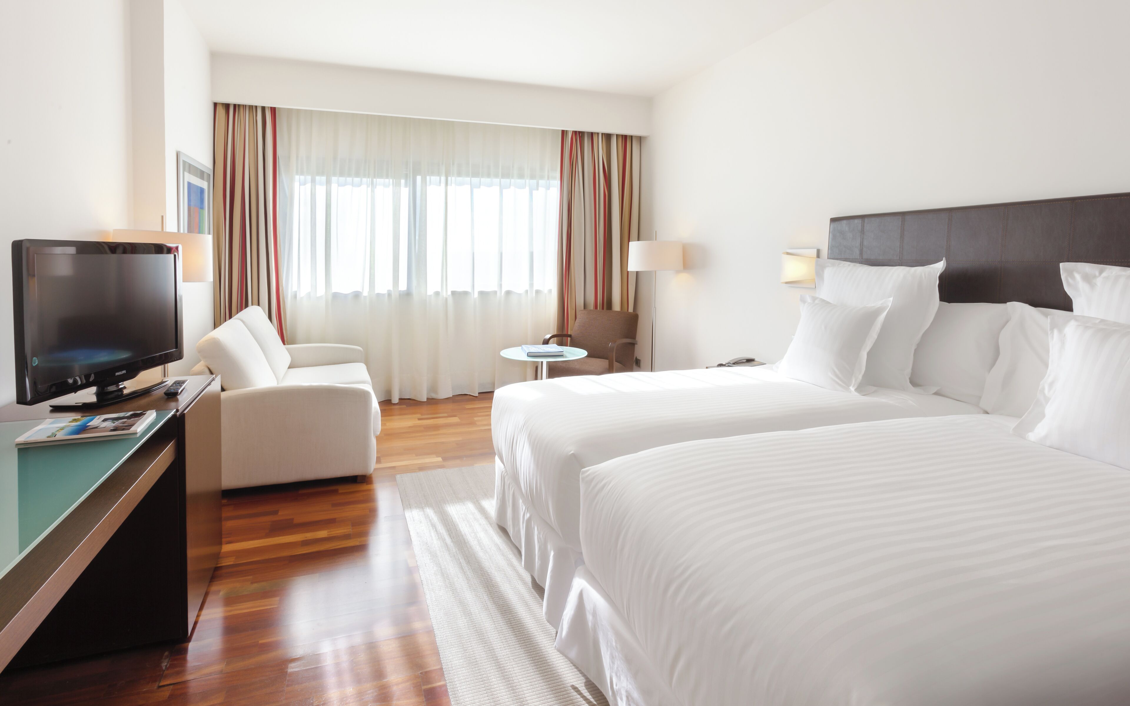 A hotel room with a bed, a desk with a TV, a chair, a small table, and a window with curtains.