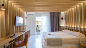 A modern hotel room with a wooden ceiling, a desk with a laptop, a comfortable chair, a bed with a patterned throw, and a view of the outside. - Bourtzi Hotel (Skiathos)