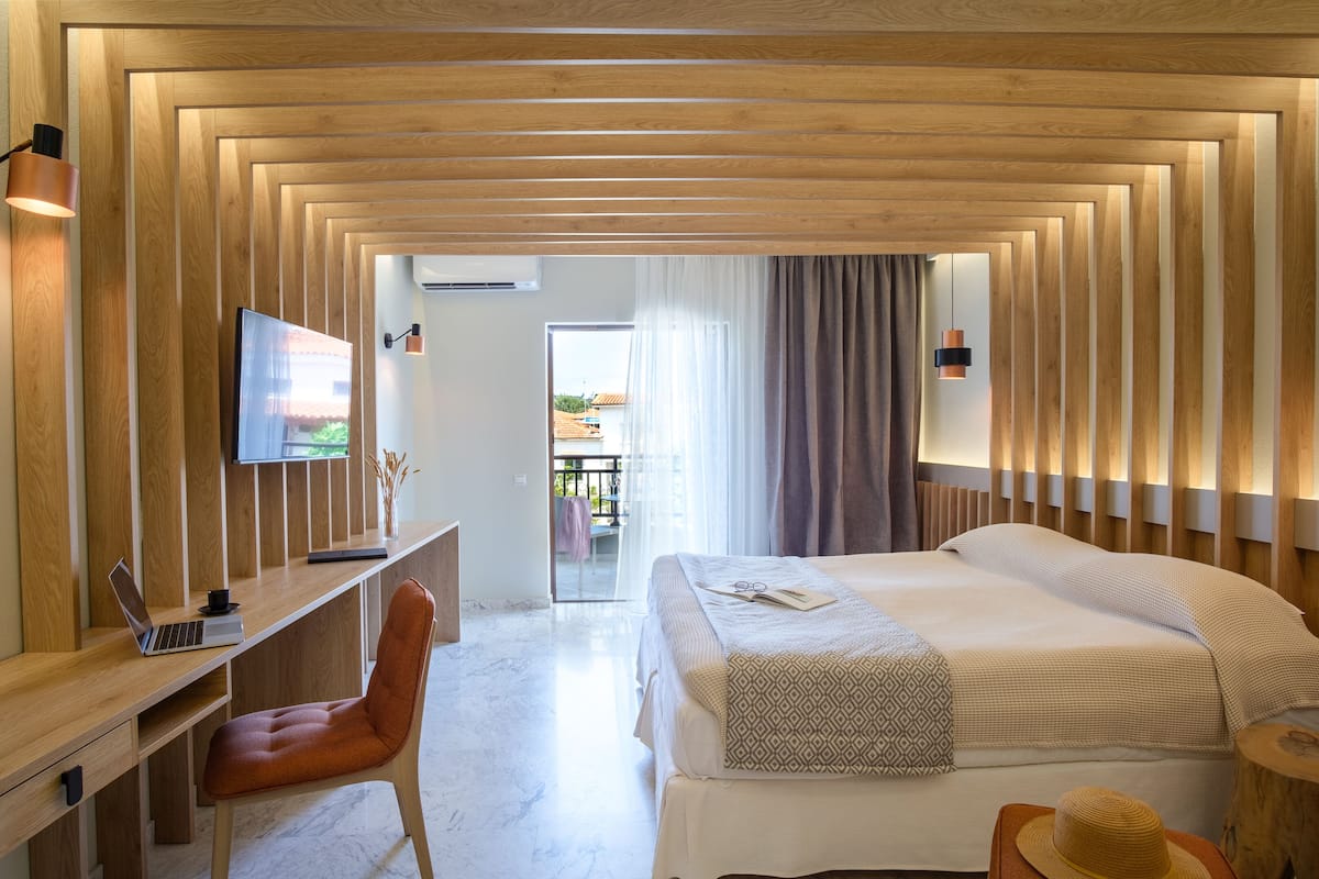 A modern hotel room with a wooden ceiling, a desk with a laptop, a comfortable chair, a bed with a patterned throw, and a view of the outside.