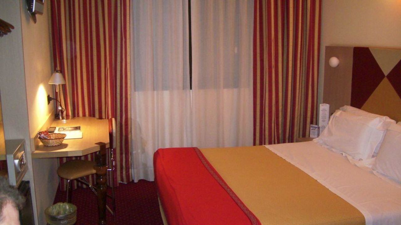 A hotel room with a bed, a desk with a lamp, a chair, and a small table with a basket.