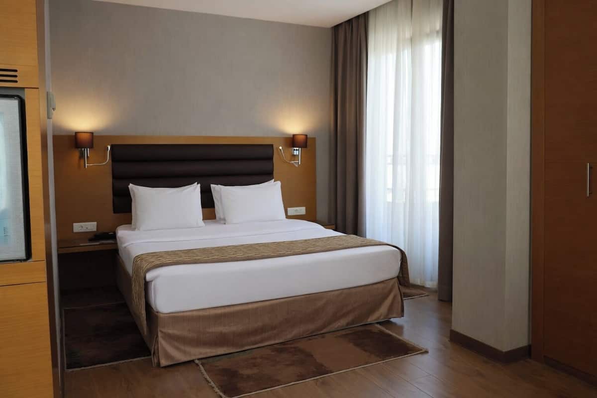 Ein modernes Hotelzimmer mit einem großen Bett, einem Holz-Kopfteil, zwei Nachttischlampen und Blick ins Freie durch transparente Vorhänge.