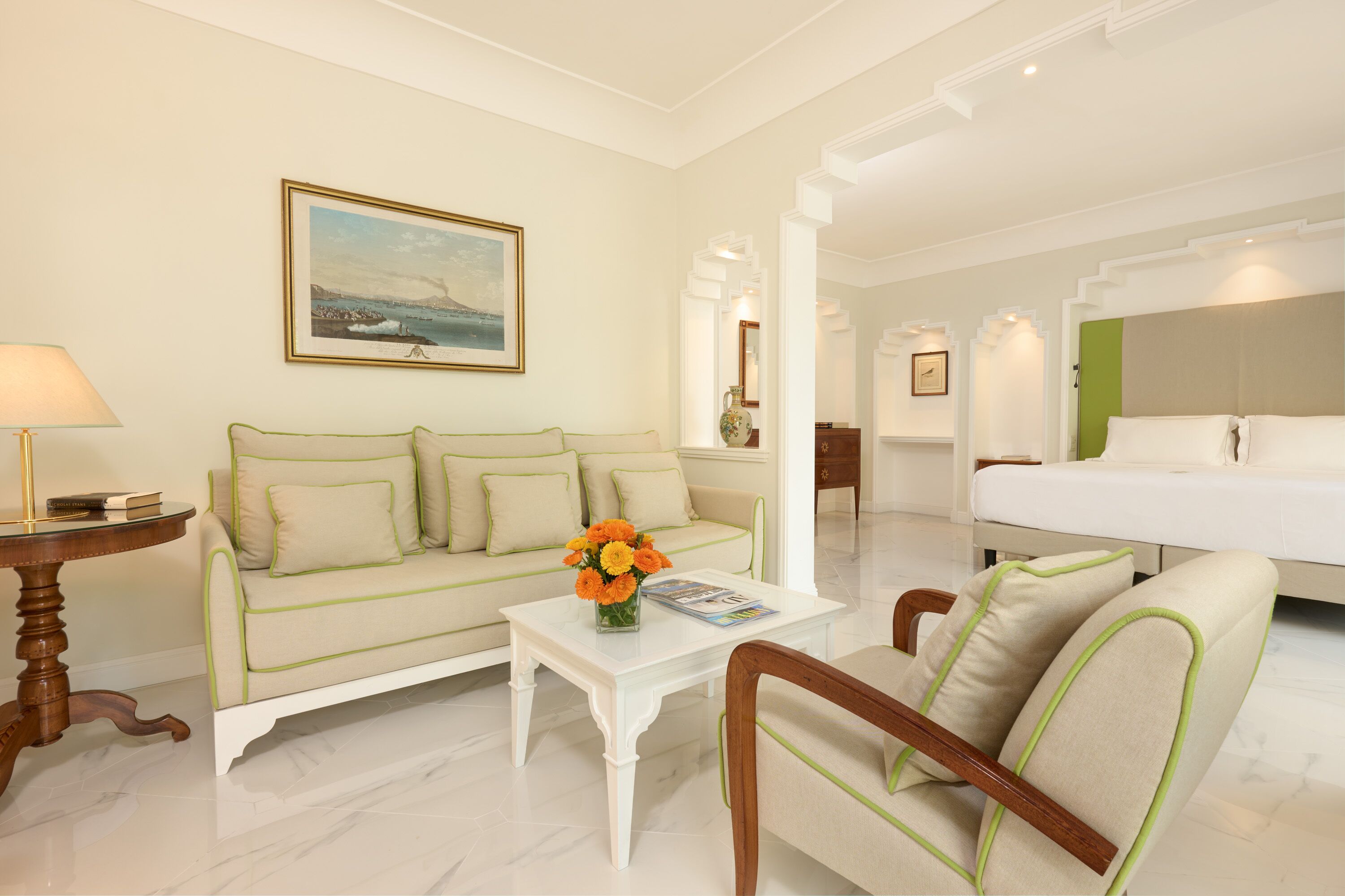 A hotel room with a sofa, a coffee table with flowers, a bed, and a painting on the wall.