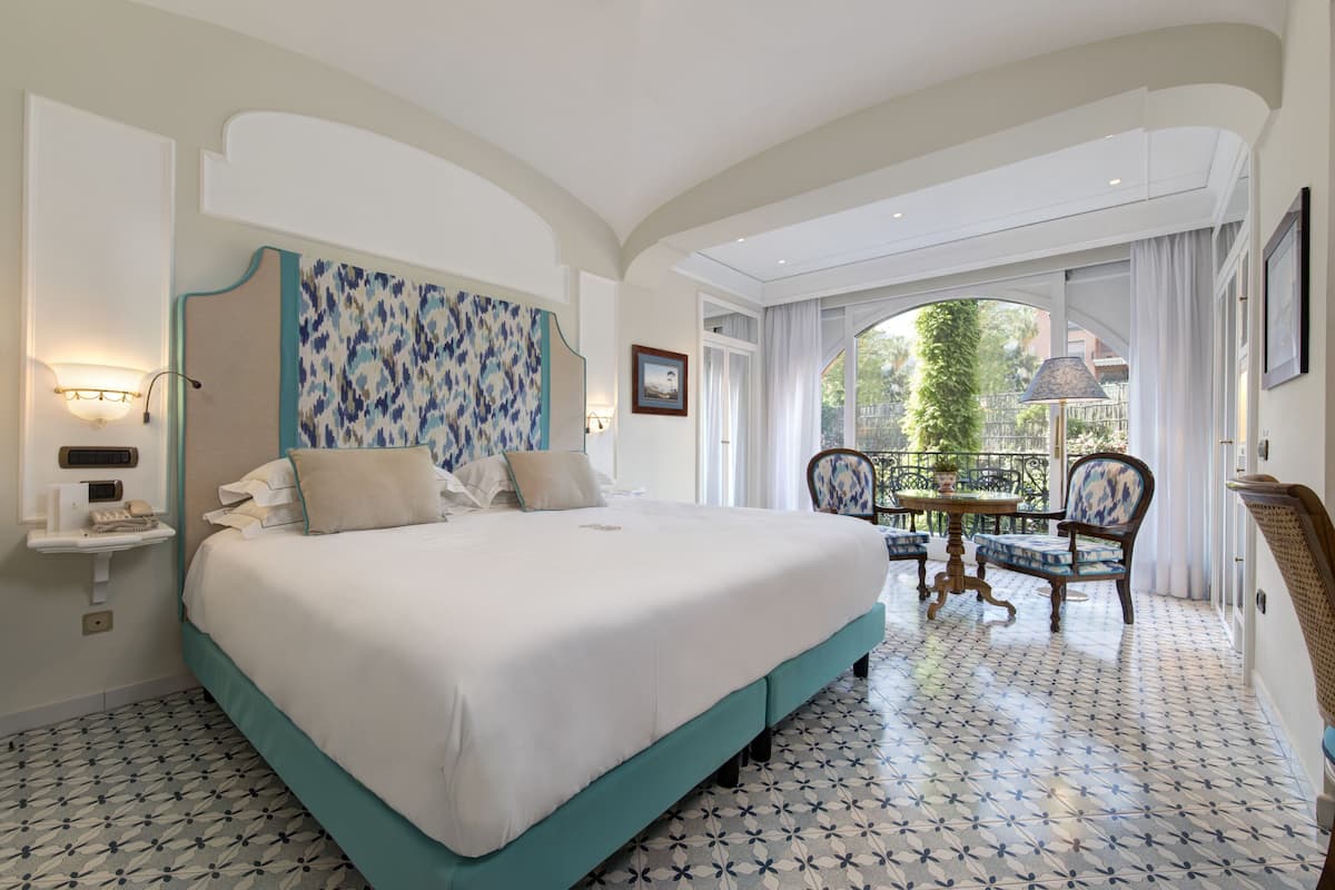 Un dormitorio amplio con una cama grande, una mesita con sillas y vistas al jardín a través de un ventanal.