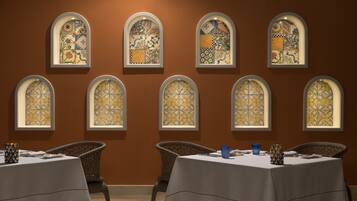 A dining area with tables set for two, each with a white tablecloth and a blue glass. The walls are adorned with arched windows featuring intricate tile patterns.