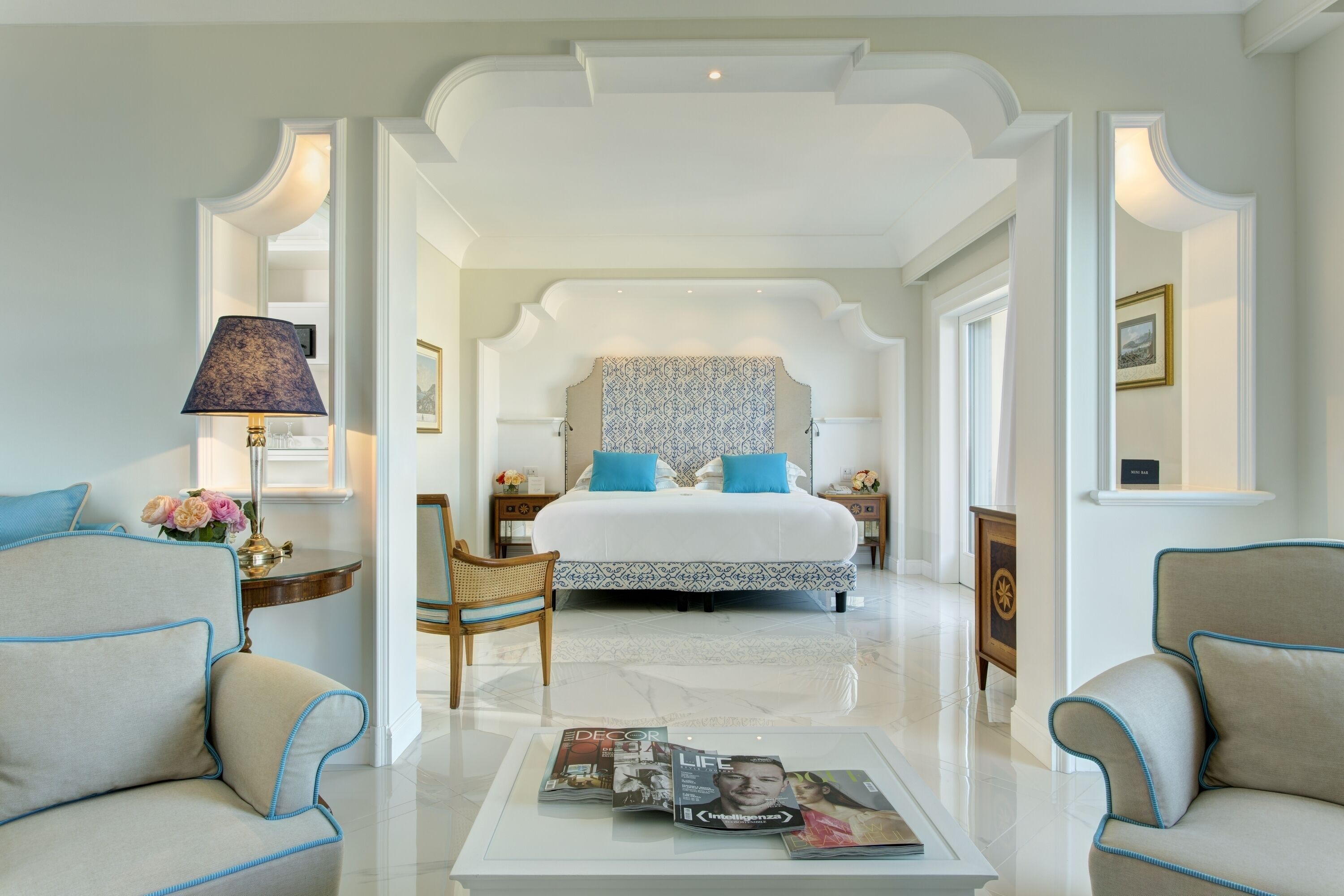 Habitación de hotel con una cama grande, dos sillones, una mesita de centro con revistas y una lámpara.