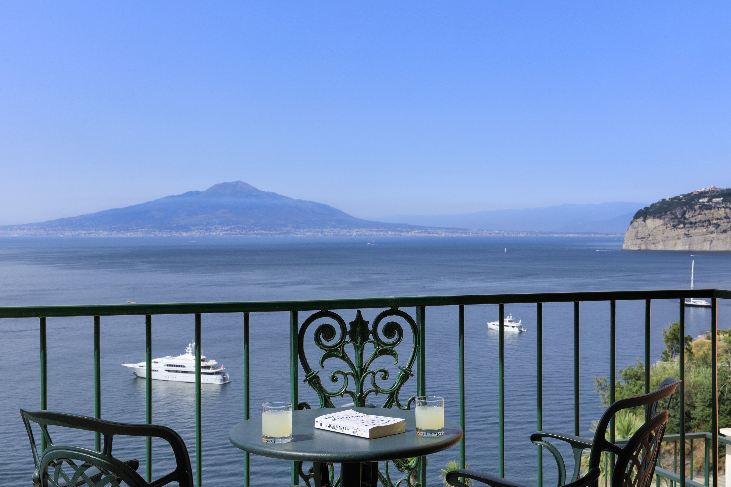 Ein Balkon mit Meerblick, ein Stuhl, ein Tisch mit einem Buch und Gläsern sowie eine ferne Berglandschaft.