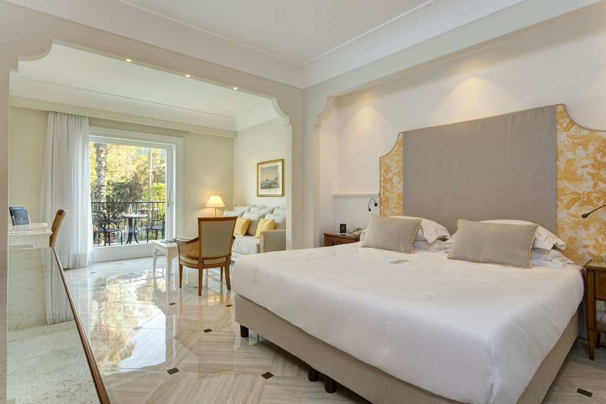 Amplia habitación de hotel con una cama grande, zona de estar con una silla y mesa, balcón con vistas a zonas verdes y un panel decorativo en la pared.