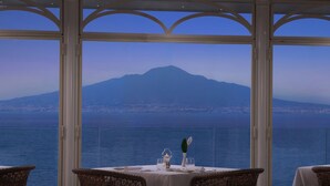 A dining area with a table set for two, overlooking a body of water and a mountain. - Grand Hotel Royal (Sorrento)