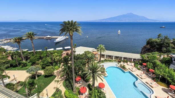 A resort with a swimming pool, palm trees, and a view of the sea and a mountain. - Grand Hotel Royal (Sorrento)