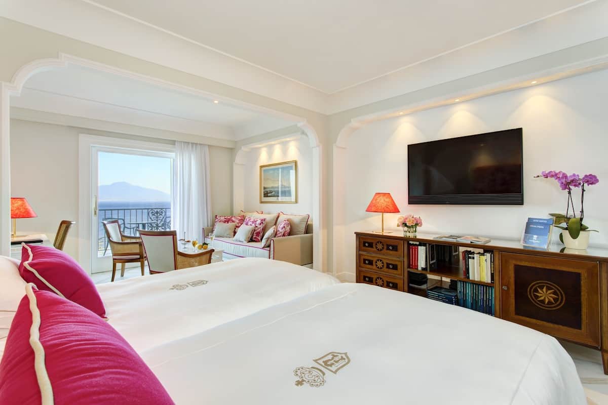 Habitación de hotel con una cama grande, televisión, balcón con vistas, escritorio con lámpara y estantería.