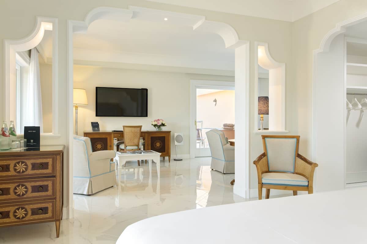 Habitación de hotel con una cama grande, un televisor de pantalla plana, un aparador de madera, un sillón blanco, una silla de mimbre y una mesita de noche con lámpara.