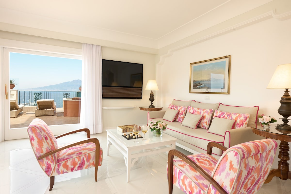 A hotel room with a balcony, a sofa, two armchairs, a coffee table, and a TV.