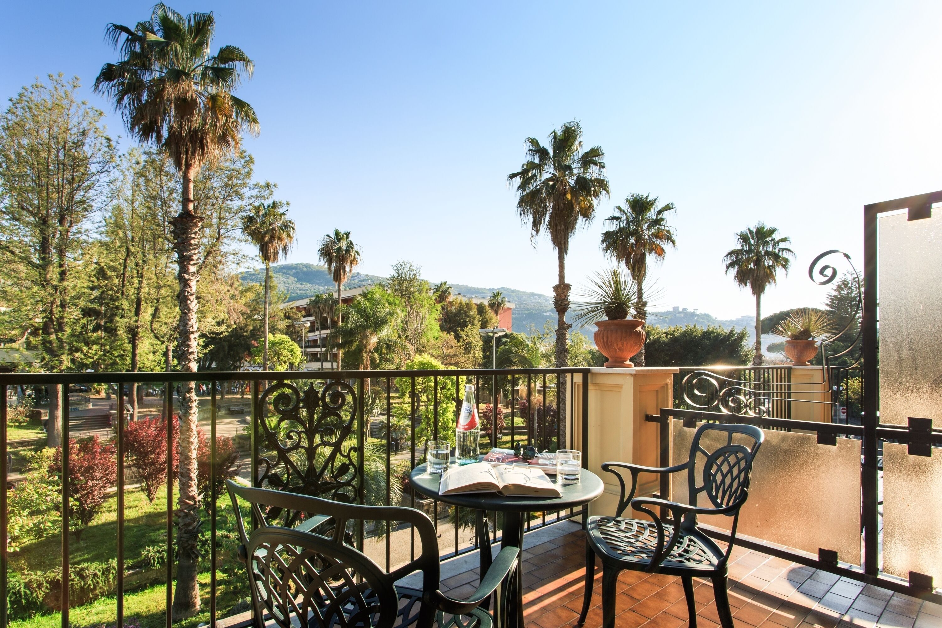Un balcone con tavolo e sedie, affacciato su un giardino con palme e vista su colline lontane.
