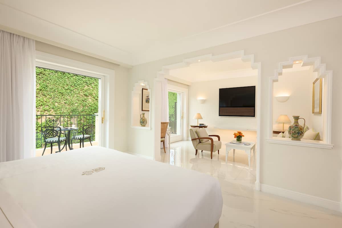 Habitación de hotel con una cama grande, balcón con sillas, televisor montado en la pared y una mesita de noche con lámpara y florero.