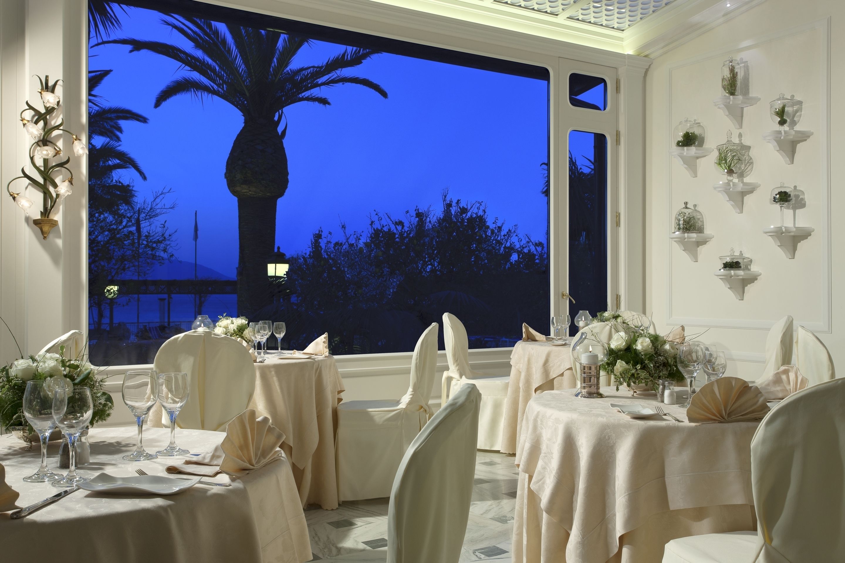 Un comedor con mesas dispuestas para una comida, sillas y un amplio ventanal con vistas a palmeras y un cielo nocturno.