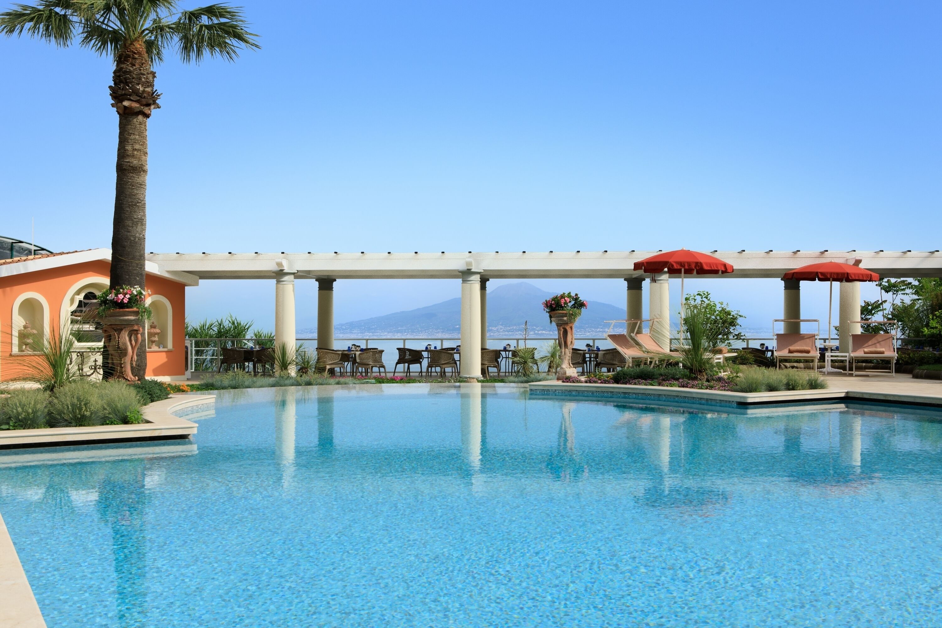 A pool area with lounge chairs and umbrellas, a palm tree, and a view of a mountain.